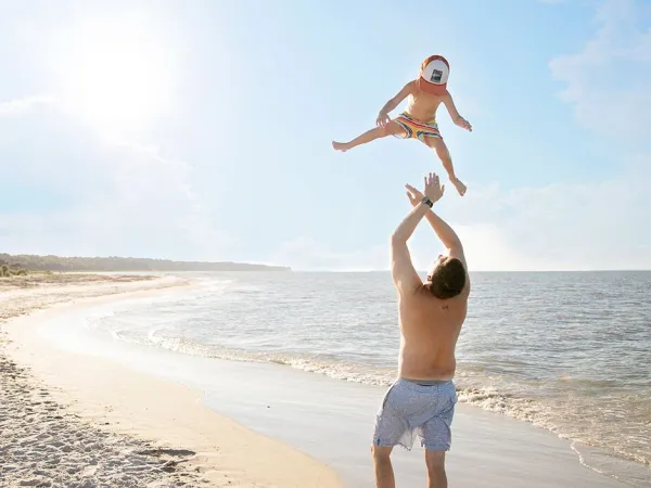 Un père lance son petit garçon en l'air sur la plage du camping de Roan Sole Family Camping Village.